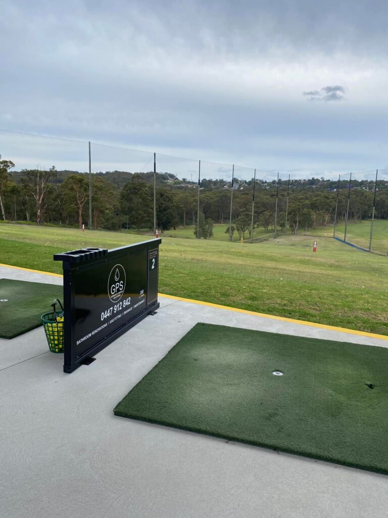 A outdoor golf driving range bay with a synthetic grass mat and a black divider featuring a "GPS" plumbing company advertisement and a basket of yellow golf balls.