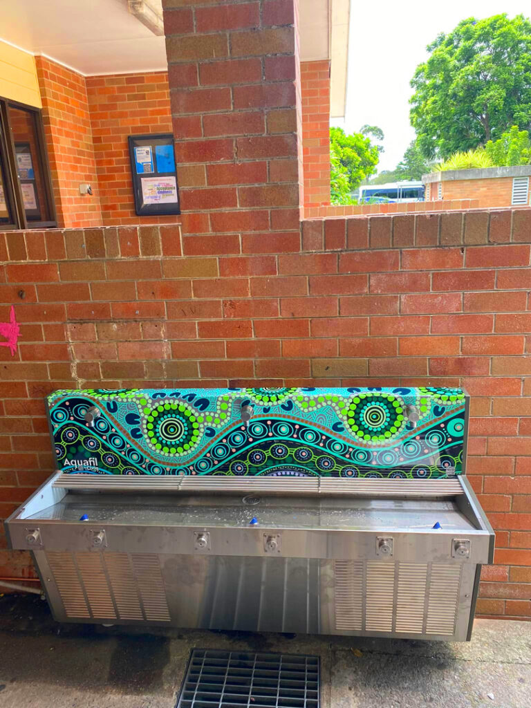 An Aquafil outdoor drinking fountain and bottle refill station featuring a vibrant Indigenous dot art design in green and blue, mounted against a red brick wall.