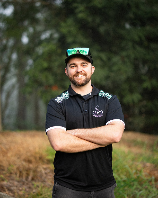 A smiling man with a beard posing with his arms crossed outdoors, wearing a black Gordon Plumbing Solutions (GPS) polo shirt and a black baseball cap with green reflective sunglasses perched on top.