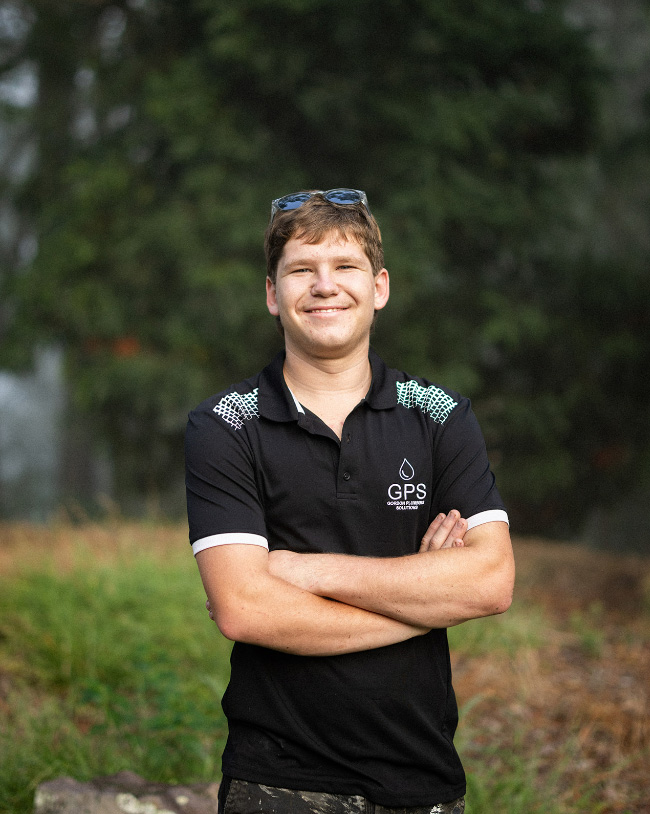 A smiling young man posing with his arms crossed outdoors, wearing a black Gordon Plumbing Solutions (GPS) polo shirt with sunglasses perched on his head.