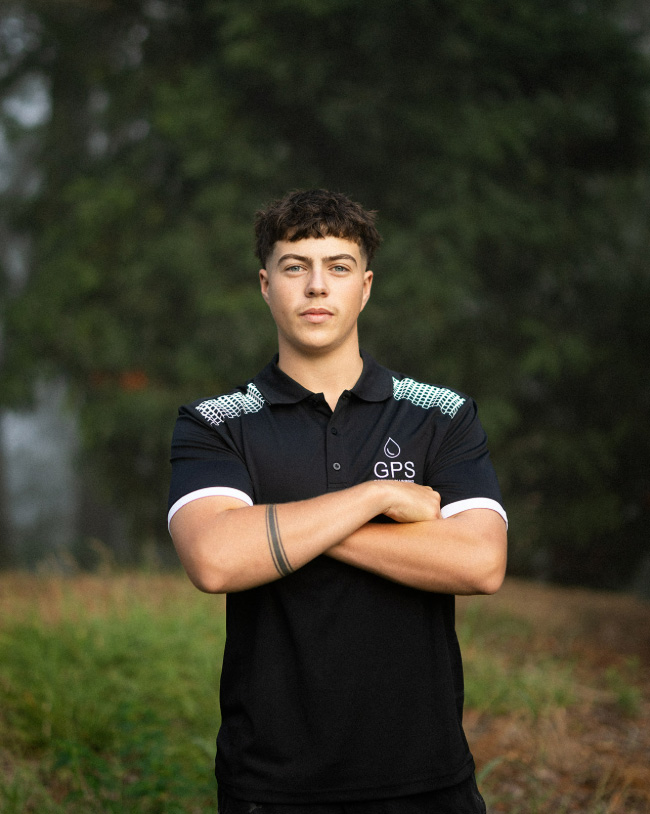 A young man with dark hair posing with his arms crossed outdoors, wearing a black Gordon Plumbing Solutions (GPS) polo shirt with a forearm tattoo visible.