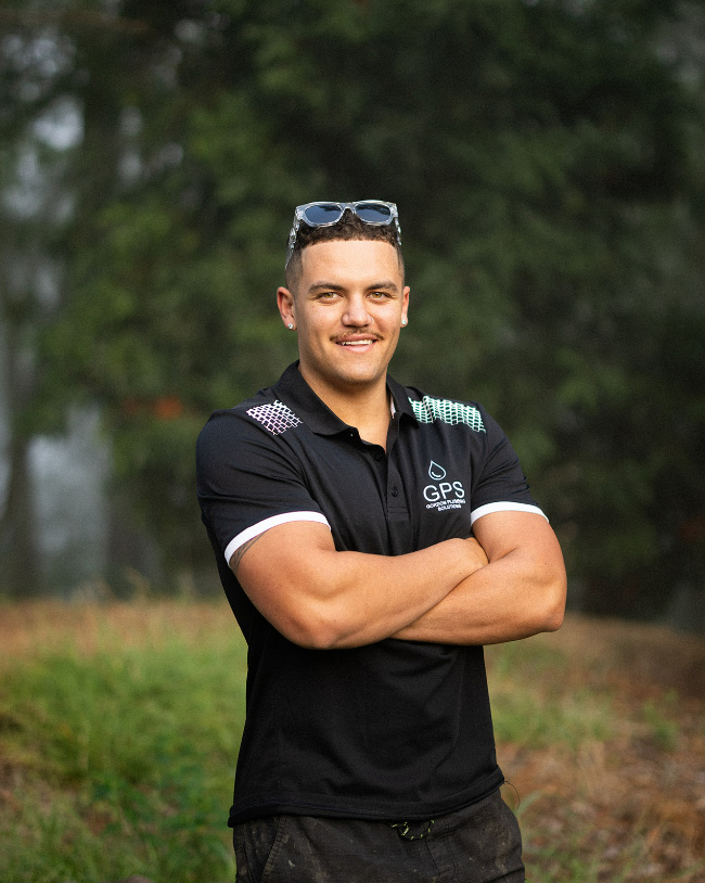 A smiling man posing with his arms crossed outdoors, wearing a black Gordon Plumbing Solutions (GPS) polo shirt with clear-framed sunglasses on his head.