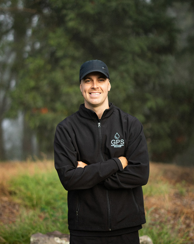 A smiling man posing with his arms crossed outdoors, wearing a black Gordon Plumbing Solutions (GPS) zip-up jacket and a black baseball cap.