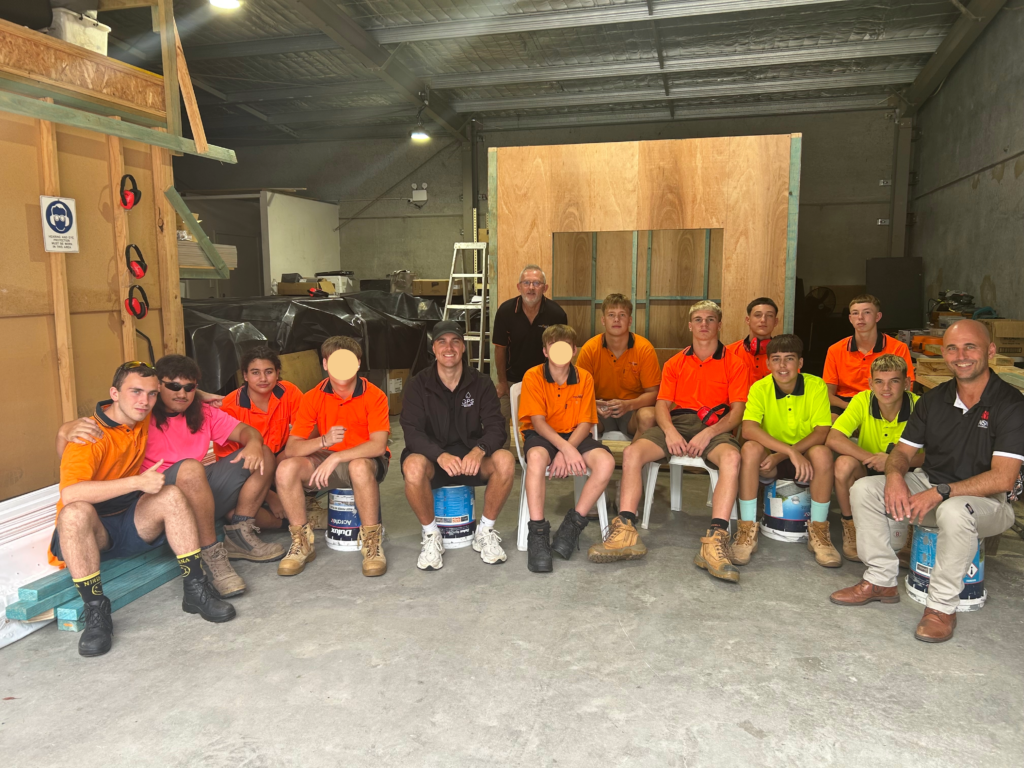 A group of trade students and instructors posing for a photo inside a workshop, with most students wearing high-visibility orange and yellow work shirts while seated on buckets and chairs in front of a wooden project structure.