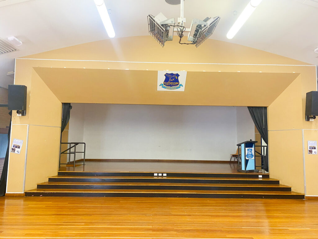 An empty school stage at Glendore Public School, featuring polished wooden floors, several wide steps leading up to the platform, and a blue podium on the right side.