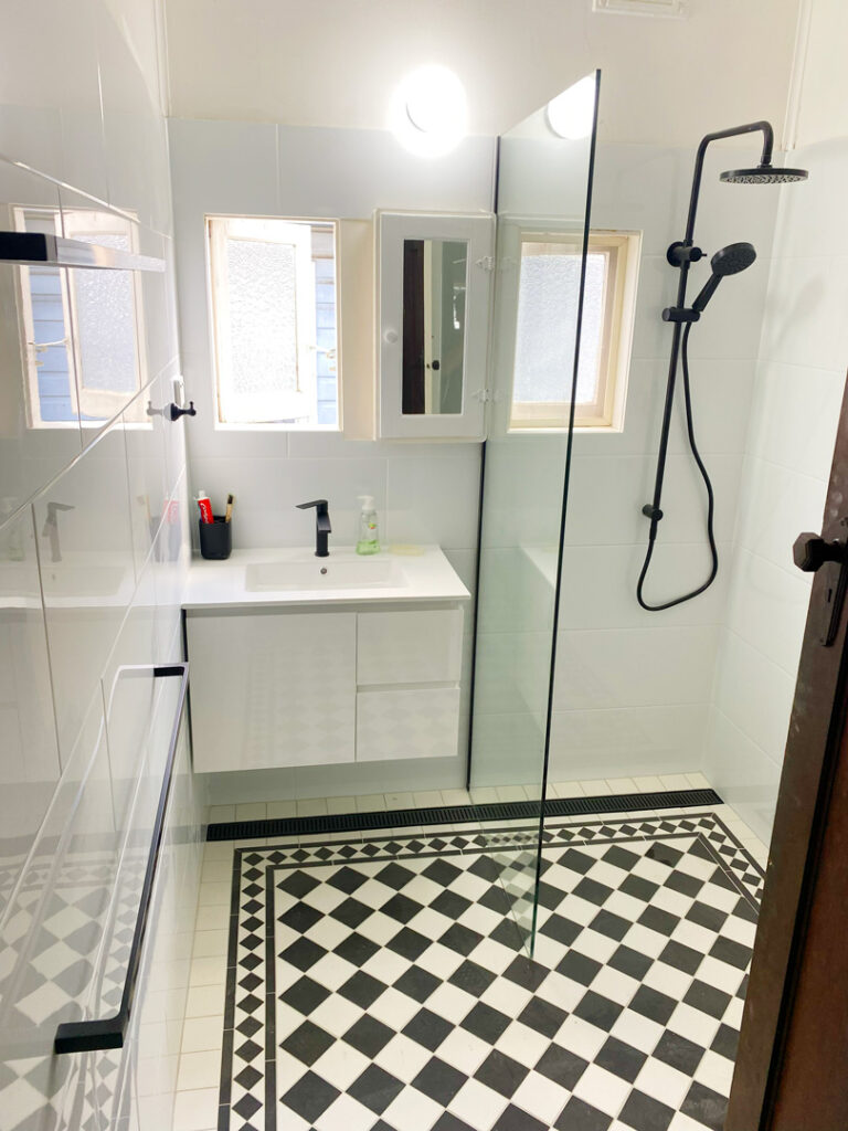 A modern, renovated bathroom featuring a black-and-white chequered tile floor, a floating white vanity with a black faucet, and a walk-in shower with a glass partition and black rainfall showerhead.