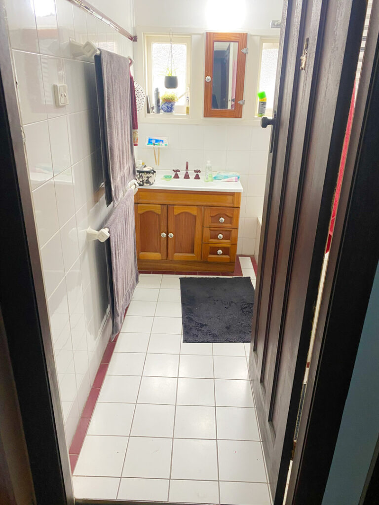 A view through a wooden doorway into an older bathroom with white floor and wall tiles, featuring a timber vanity with reddish-brown taps and purple towels hanging on the wall.